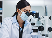 eine Frau im Laborkittel und mit Mundschutz schaut in ein Mikroskop