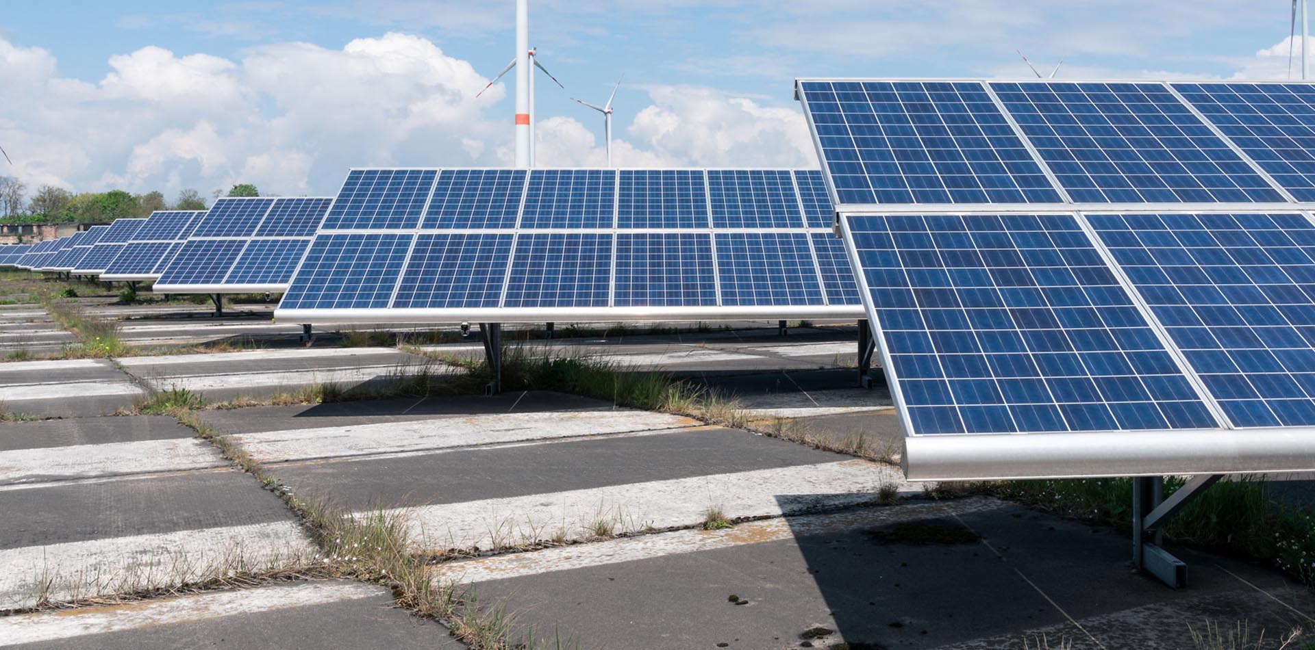Photovoltaik-Paneele stehen auf Betonplatten, im Hintergund Windräder erkennbar